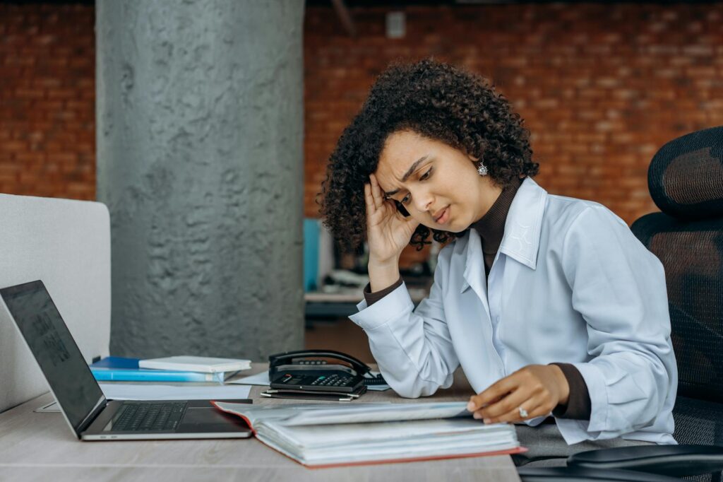 Exhausted woman of color sitting at a desk, holding her head in one hand while reading through documents, surrounded by a laptop and office supplies, indicating stress and burnout.