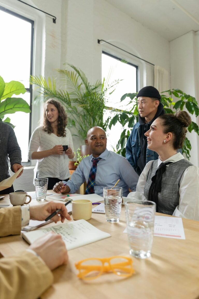 A diverse group of professionals, including people of color and LGBTQ+ individuals, smiling and collaborating around a meeting table in a bright, plant-filled office space.