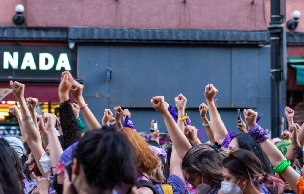 A group of diverse individuals with raised fists in solidarity, wearing purple, green, and other colored bandanas, participating in a public demonstration.