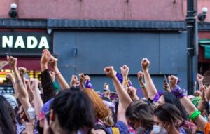A group of diverse individuals with raised fists in solidarity, wearing purple, green, and other colored bandanas, participating in a public demonstration.