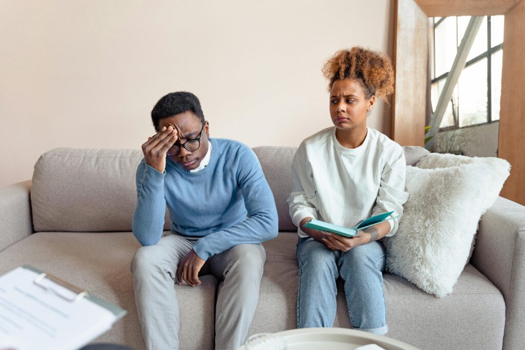 Stressed couple sitting apart on couch during relationship conflict, man holding head while woman looks away with concern