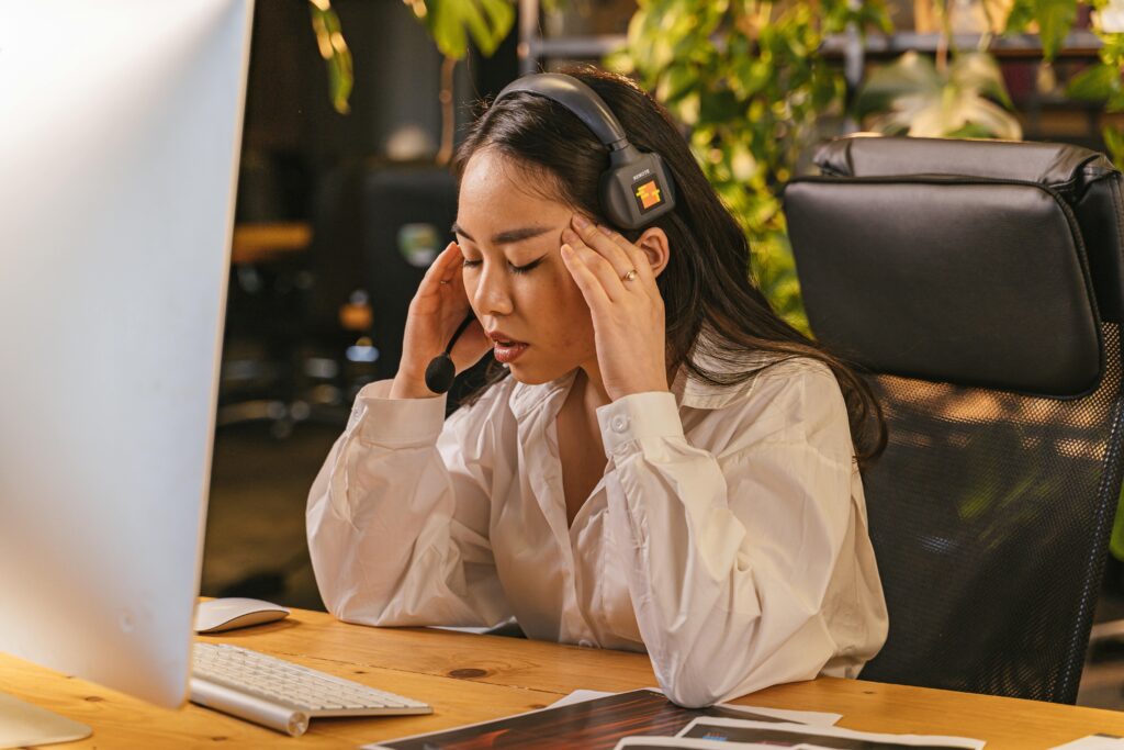 A young woman wearing a headset sits at a desk with her eyes closed, holding her temples, appearing stressed while working on a computer.