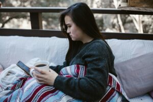 Woman sitting on a porch wrapped in a blanket, holding coffee and looking at her phone thoughtfully, reflecting on feelings about a romantic interest.